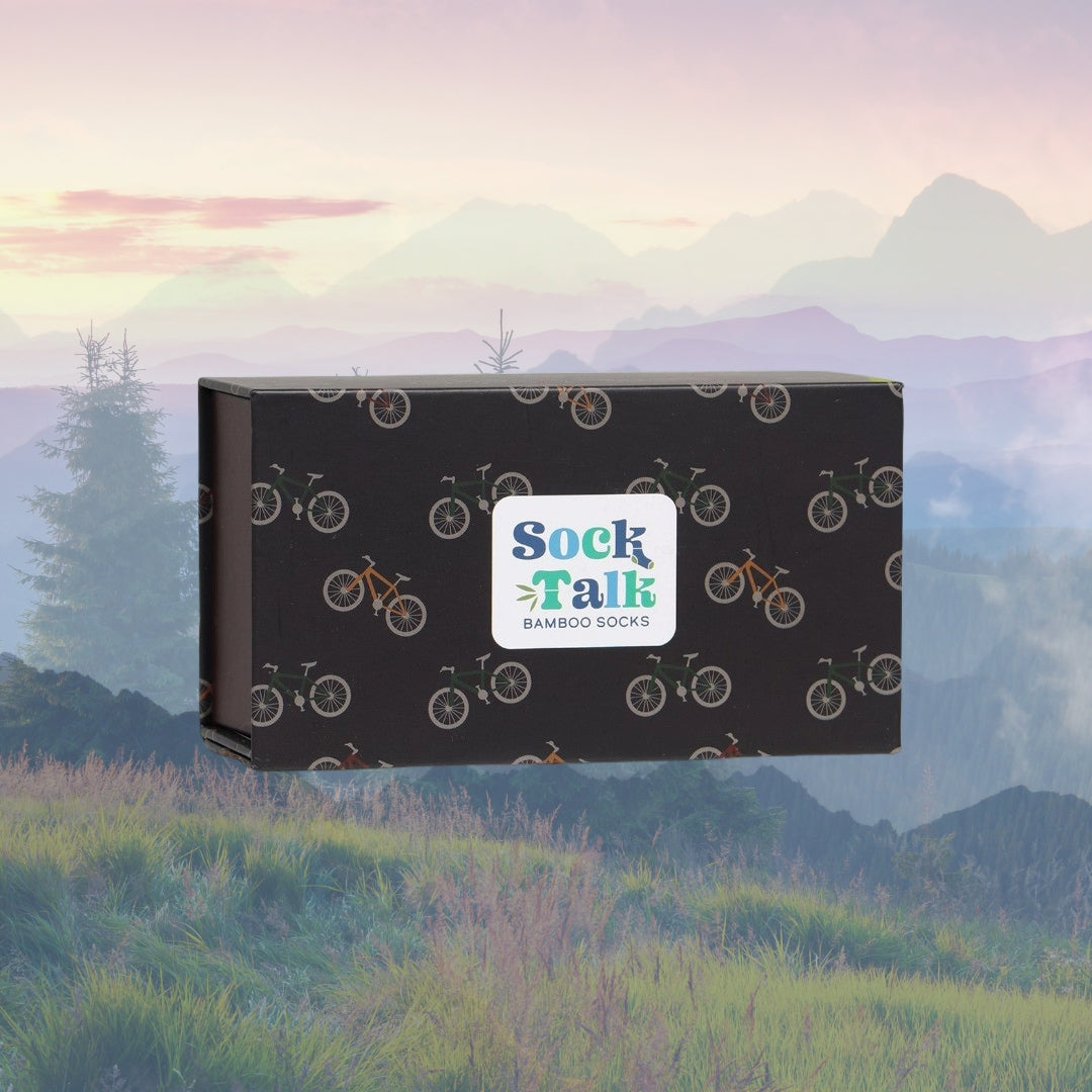 En lukket gaveeske fra Sock Talk med et mønster av sykler på svart bakgrunn. Esken har en hvit logo med teksten "Sock Talk Bamboo Socks". Bakgrunnen viser et rolig landskap med fjell og trær i pastellfarger.