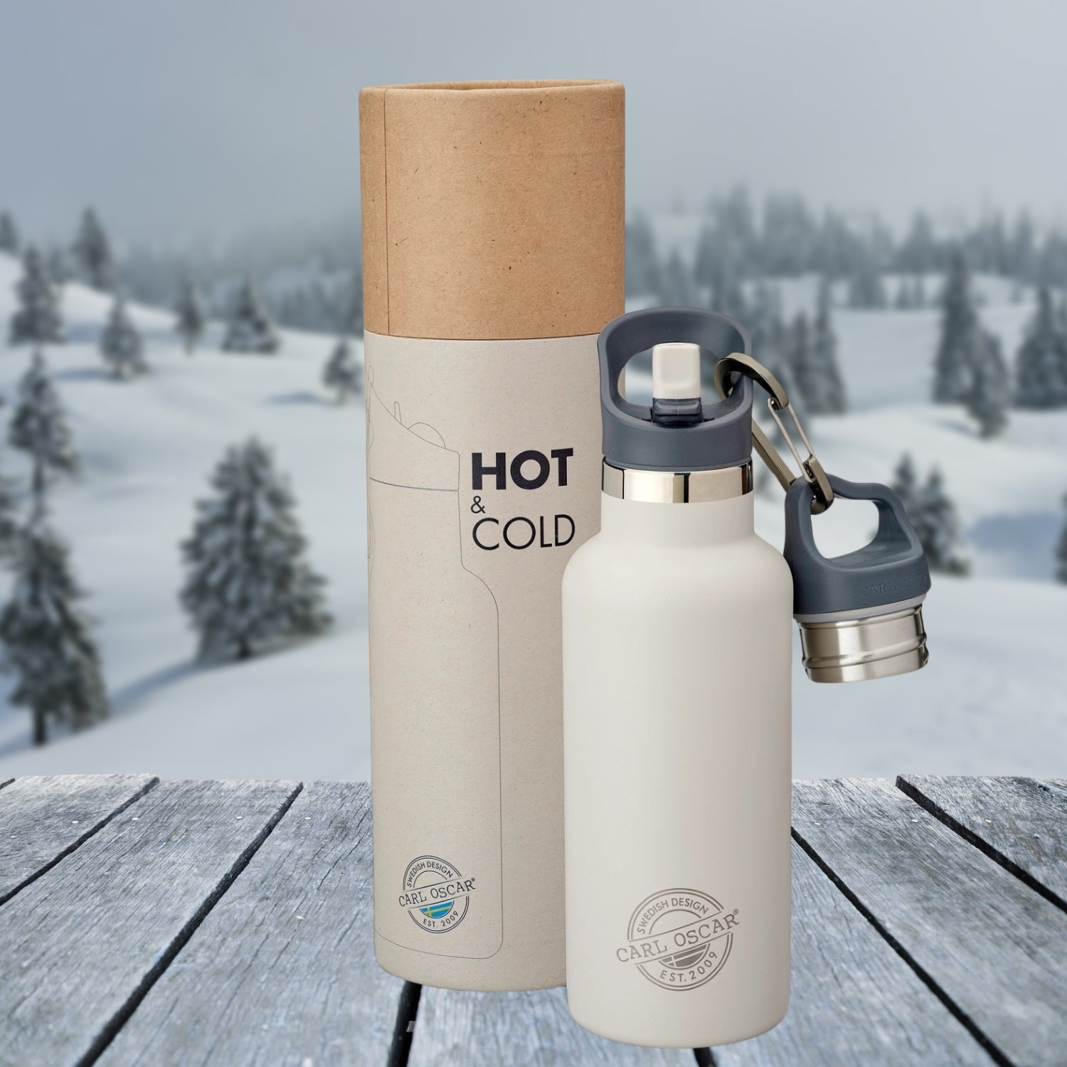 En greige Carl Oscar termoflaske med sølvfarget lokk og grå bærehåndtak, plassert foran en sylinderformet gaveeske med teksten "HOT & COLD." Flasken har et minimalistisk design med Carl Oscar-logo gravert nederst. Bakgrunnen viser et vinterlandskap med snødekte trær og åser, mens flasken står på en trebordsoverflate med frostet utseende.