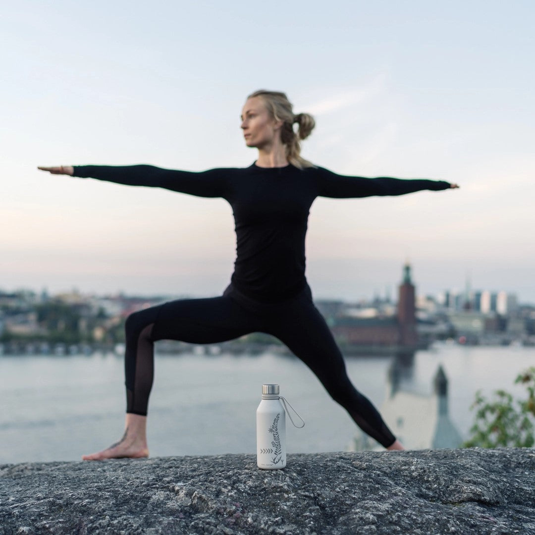 En kvinne i svart treningsklær utfører en yogaøvelse på en stein med en beige Carl Oscar termoflaske plassert foran henne. Bakgrunnen viser en rolig byutsikt med vann og bygninger.