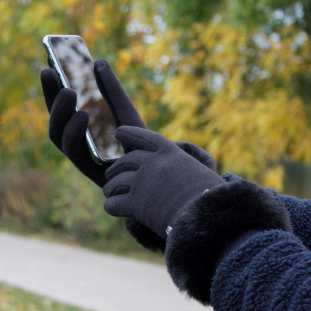 Et par svarte hansker med myk fuskepels på mansjettene, som brukes til å betjene en smarttelefon. En person iført en mørk jakke står utendørs med høstfargede trær i bakgrunnen. Hanskene har en touchfunksjon for bruk med skjermer.